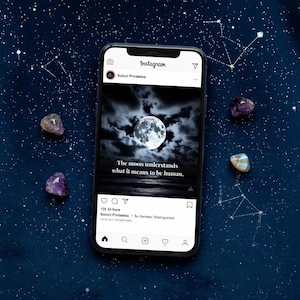 May include: A smartphone displays an Instagram post with a celestial theme. The post features a full moon in a cloudy sky with the text "The moon understands what it means to be human." Amethyst and other crystals are scattered around the phone on a dark blue surface.
