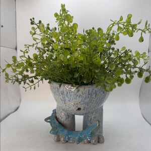May include: A ceramic planter with a speckled light blue and white glaze, shaped like a creature with two thick legs and a spiked collar around its base. The planter is filled with a lush, vibrant green artificial plant with small, rounded leaves.