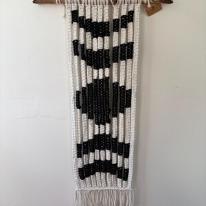 May include: A macrame wall hanging with a geometric design. The piece features alternating vertical stripes of white and black, with a fringe at the bottom. It is suspended from a natural wooden branch.