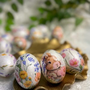 May include: Decorative Easter eggs with floral and bunny designs. The eggs are white with colorful illustrations of flowers and a brown bunny. They are arranged on a gold-colored, leaf-shaped tray, suggesting a springtime theme.