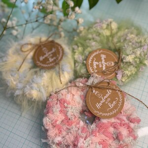 May include: Three handmade scrunchies in pastel colors: yellow, green, and pink. Each scrunchie is fluffy and textured, with a round tag that reads "Handmade with love" tied with twine. The scrunchies are arranged on a light blue surface.
