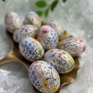 May include: A collection of decorative Easter eggs with floral designs in a gold-colored tray. The eggs feature various flowers in shades of pink, blue, yellow, and white. The tray sits on a white lace surface.