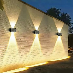 Pode incluir: Três luminárias de parede externas são montadas em uma parede branca texturizada. Cada luminária é retangular e emite um feixe de luz brilhante e focado para baixo, criando um padrão triangular. A cena se passa ao entardecer, com um céu azul escuro.