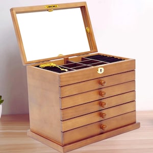 May include: A wooden jewelry box with a hinged glass top and multiple drawers. The box is made of light brown wood and has a keyhole on the front. The drawers have round wooden knobs. The interior is lined with black fabric.
