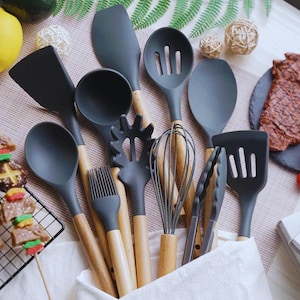 Könnte beinhalten: Eine Sammlung dunkelgrauer Küchenutensilien mit natürlichen Holzgriffen. Das Set umfasst Spatel, Löffel, einen Schneebesen, Zangen und einen Backpinsel. Die Utensilien sind in einem weißen Behälter angeordnet.