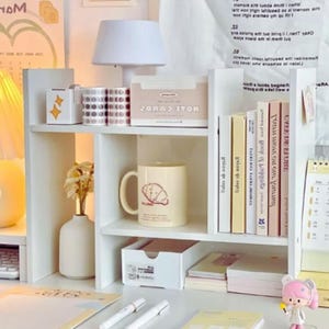 May include: A white desk organizer with multiple shelves, holding a small lamp, a mug with a croissant design, books, and stationery. The organizer also has a small vase with flowers and a box with the text "HOLE COBD2".