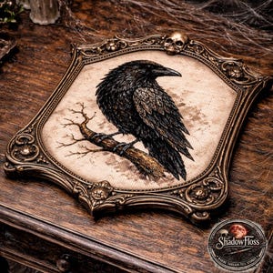 May include: A framed cross-stitch of a black raven perched on a branch. The artwork is set in an ornate, antique-style gold frame with a skull detail. The piece is displayed on a wooden surface, with candles and cobwebs in the background.