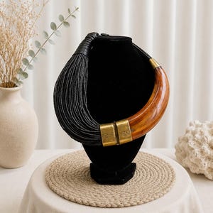 May include: A statement necklace featuring a large, curved, polished brown horn-like element connected to a thick bundle of thin black cords. The piece is accented with two cylindrical brass metal connectors that feature subtle etched line details.