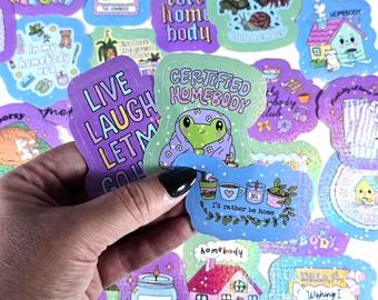Może przedstawiać: Kolekcja kolorowych naklejek z różnymi wzorami i tekstami. Jedna naklejka mówi "Certified Homebody" z ilustracją żaby. Inne naklejki zawierają frazy takie jak "Live Laugh Let Go" i "I'd rather be home."