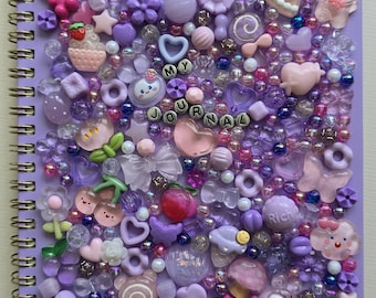 Może przedstawiać: Notatnik spiralny z jasnofioletową okładką, gęsto ozdobioną różnorodnymi fioletowymi, różowymi i przezroczystymi plastikowymi koralikami, zawieszkami i kaboszonami w kształcie serc, kwiatów, motyli i jedzenia, z napisem MY JOURNAL z koralików.