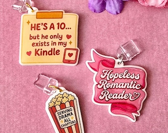 Może przedstawiać: Trzy akrylowe zawieszki do urządzeń z nadrukowanymi wzorami. Jedna to żółta karteczka samoprzylepna z napisem HE'S A 10... but he only exists in my Kindle. Druga to różowa wstążka z napisem Hopeless Romantic Reader. Trzecia to kubełek popcornu z napisem SERVING DRAMA ALL CHAPTERS.