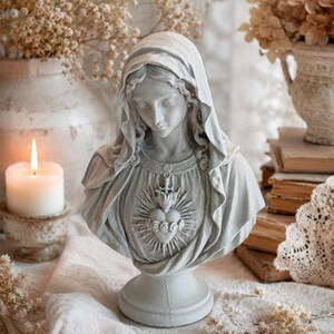 May include: A light gray stone-colored bust of a figure wearing a draped head covering and a garment featuring a central emblem of a heart surrounded by rays of light and topped with a small cross, set on a circular pedestal base.