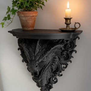 Pode incluir: Uma prateleira de parede preta com uma escultura detalhada de cabeça de dragão como suporte. A superfície superior plana contém um vaso de terracota com uma planta de hera verde e um pequeno castiçal de latão com uma vela branca acesa.