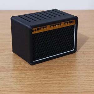 Può includere: Un amplificatore per chitarra nero in miniatura con una griglia per altoparlanti strutturata e un pannello di controllo color oro. L'amplificatore ha un bordo bianco e una parte superiore a fessura. Replica dettagliata.
