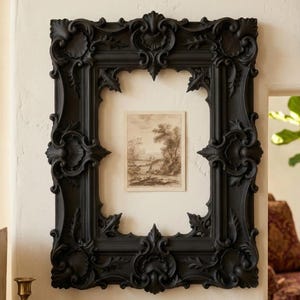 Può includere: Una grande cornice nera ornata con intricati dettagli floreali e a volute. La cornice racchiude una piccola stampa di paesaggio in seppia. La cornice è montata su una parete bianca, creando un contrasto sorprendente.