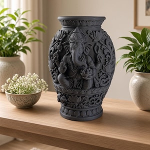 Puede incluir: Un jarrón decorativo de color negro mate que presenta un relieve detallado de la deidad Ganesha sentada en el centro. La superficie está adornada con intrincados patrones florales, motivos de loto y símbolos tradicionales alrededor.