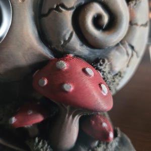 May include: A three-dimensional sculpture featuring a cluster of red mushrooms with white spots at the base of a large, textured, spiral-shaped object. The mushrooms have visible horizontal layer lines and a matte finish, set against a weathered, greyish-brown background.