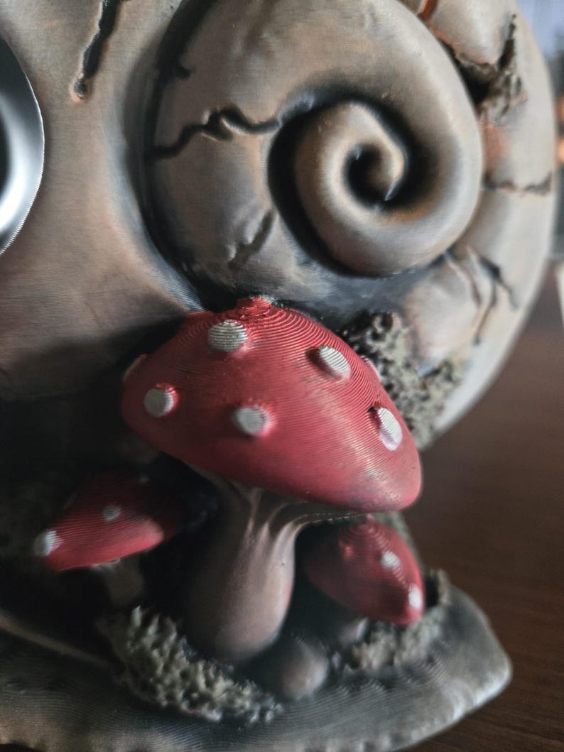 May include: A three-dimensional sculpture featuring a cluster of red mushrooms with white spots at the base of a large, textured, spiral-shaped object. The mushrooms have visible horizontal layer lines and a matte finish, set against a weathered, greyish-brown background.