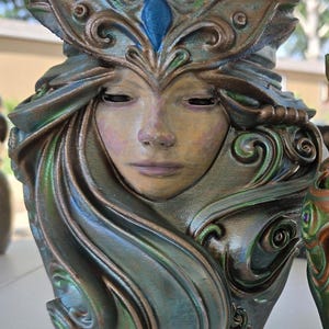 May include: A sculpted vessel featuring a human face with closed eyes, surrounded by flowing, ornate hair patterns. The sculpture is finished in a metallic bronze and green patina with a single blue teardrop-shaped accent on the forehead.