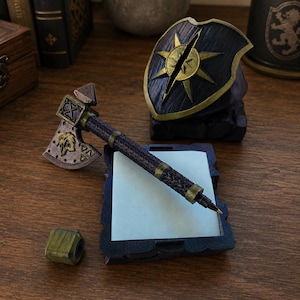 May include: A dark blue and gold pen shaped like a battle axe, resting on a rectangular note holder with light blue paper. A matching dark blue and gold shield-shaped pen stand sits behind the note holder, accompanied by a small green cube.