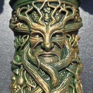 May include: A cylindrical container featuring a relief sculpture of a face integrated with tree branches and roots. The object is finished in a deep forest green color with metallic gold highlights applied to the raised details of the design.
