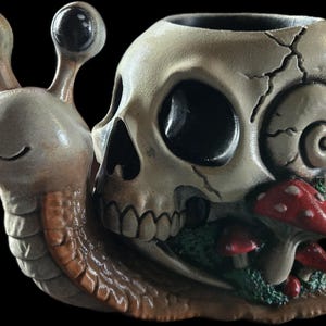 May include: A snail-shaped planter featuring a human skull as the shell. The snail has a light brown body with two prominent eyes on stalks. The skull shell is off-white with cracks and is decorated with small red mushrooms and green moss accents.