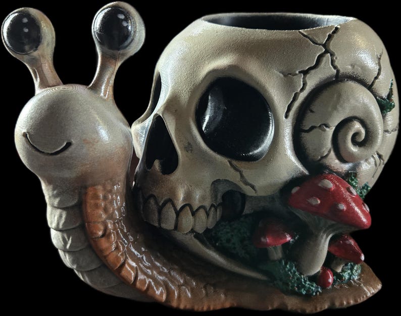 May include: A snail-shaped planter featuring a human skull as the shell. The snail has a light brown body with two prominent eyes on stalks. The skull shell is off-white with cracks and is decorated with small red mushrooms and green moss accents.