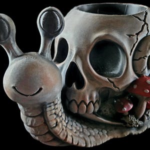May include: A grey, snail-shaped planter featuring a human skull shell with a cracked surface. The snail has a smiling face and two eye stalks. Small red mushrooms with white spots are attached to the side of the skull, near the base.