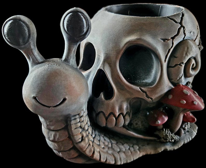 May include: A grey, snail-shaped planter featuring a human skull shell with a cracked surface. The snail has a smiling face and two eye stalks. Small red mushrooms with white spots are attached to the side of the skull, near the base.