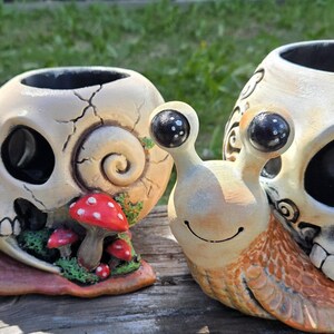 May include: Two snail-shaped planters featuring off-white skull shells with black eye sockets and teeth. The left snail includes small red mushrooms and green moss accents. Both have tan bodies with black eyes on stalks and open tops for planting.