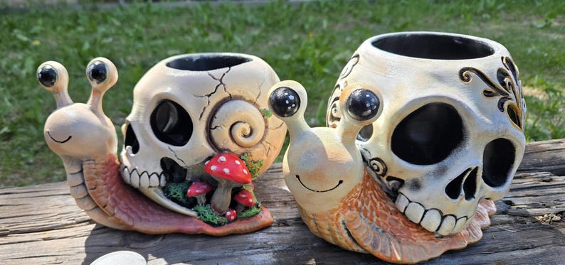 May include: Two snail-shaped planters featuring off-white skull shells with black eye sockets and teeth. The left snail includes small red mushrooms and green moss accents. Both have tan bodies with black eyes on stalks and open tops for planting.
