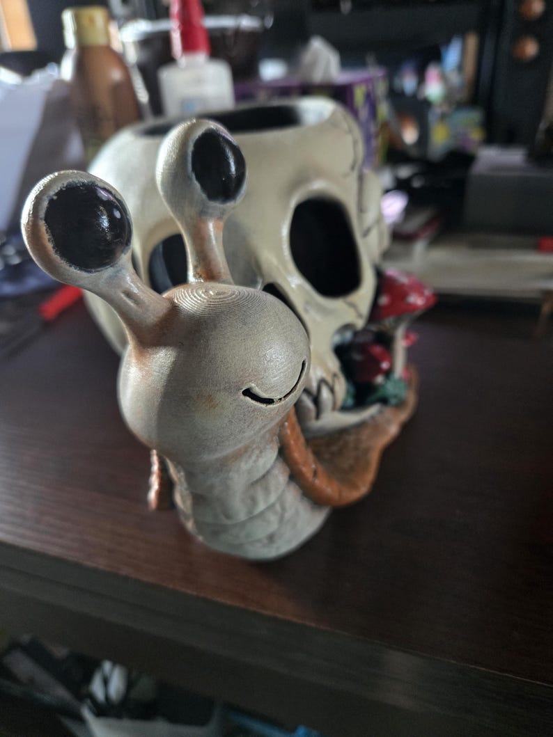 May include: A ceramic sculpture featuring a light gray snail with black eyes and a smiling mouth, positioned in front of a beige human skull. The skull is decorated with small red and white mushrooms nestled near the base of the snail's shell.