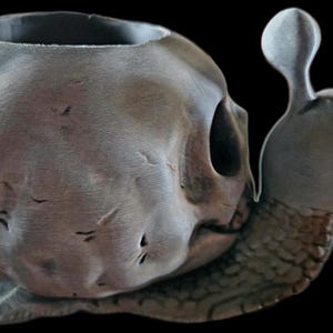 May include: A gray snail-shaped planter featuring a shell designed to resemble a human skull. The skull shell has a weathered, textured surface with visible cracks and a hollow top opening. The snail body has a detailed, segmented skin texture.