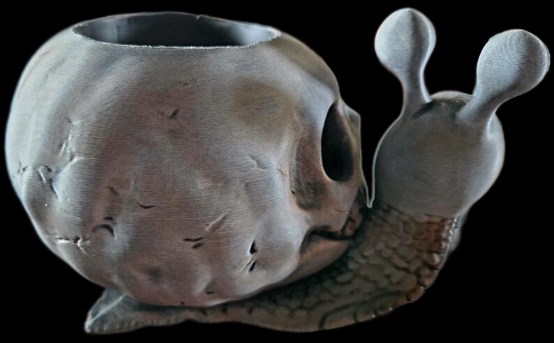 May include: A gray snail-shaped planter featuring a shell designed to resemble a human skull. The skull shell has a weathered, textured surface with visible cracks and a hollow top opening. The snail body has a detailed, segmented skin texture.
