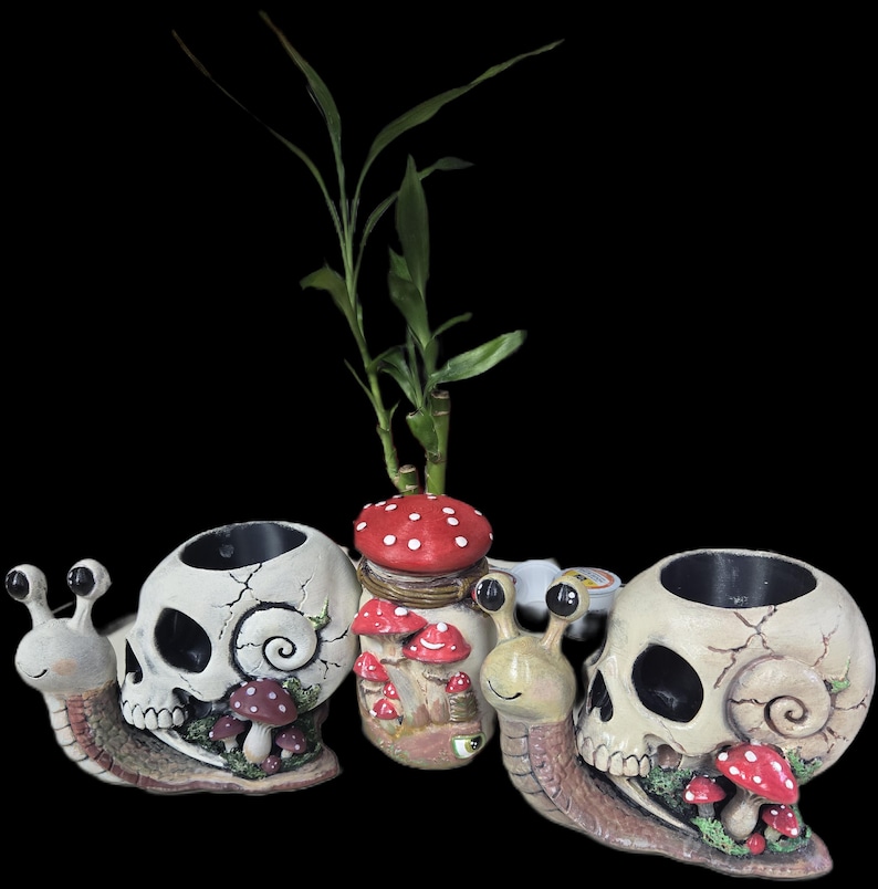 May include: Two snail-shaped planters with off-white skull shells featuring cracked textures and red mushroom accents, positioned on either side of a cylindrical container decorated with red mushrooms and topped with a green bamboo plant.