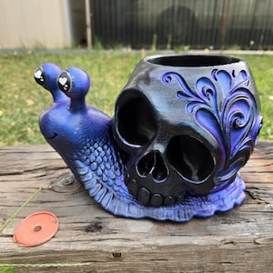 May include: A snail-shaped planter featuring a blue and purple gradient body with a textured surface and a black skull-shaped shell. The shell is decorated with carved swirling patterns and has large eye sockets and a nasal cavity. The snail has two stalks.