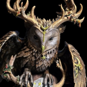 May include: A detailed figurine of an owl with large, branching antlers extending from its head. The owl features brown, textured plumage, a green beak, and a gold crescent moon symbol on its forehead. It holds a circular object with green accents.