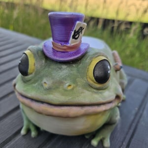 May include: A green frog figurine with large black eyes and a light-colored belly. The frog wears a purple top hat with a white tag on the side featuring the black text 10/6. The surface of the figurine shows visible 3D printed layer lines.