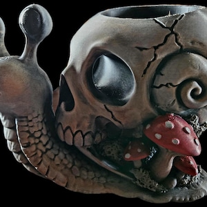 May include: A decorative snail figurine featuring a human skull as its shell, with a textured brown body and two red mushrooms with white spots growing at the base. The skull shell displays visible cracks and a hollow interior opening at the top.