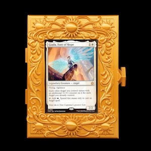 May include: A gold-colored rectangular frame with ornate sun and scrollwork detailing, housing a Magic: The Gathering trading card titled Giada, Font of Hope. The card features an angel illustration, text, and a 2/2 power and toughness rating.