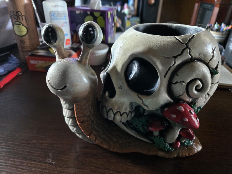 May include: A decorative snail figurine featuring a human skull as its shell. The snail has a beige body with large black eyes and a smiling mouth. The skull shell is off-white with cracks and is decorated with small red mushrooms with white spots.