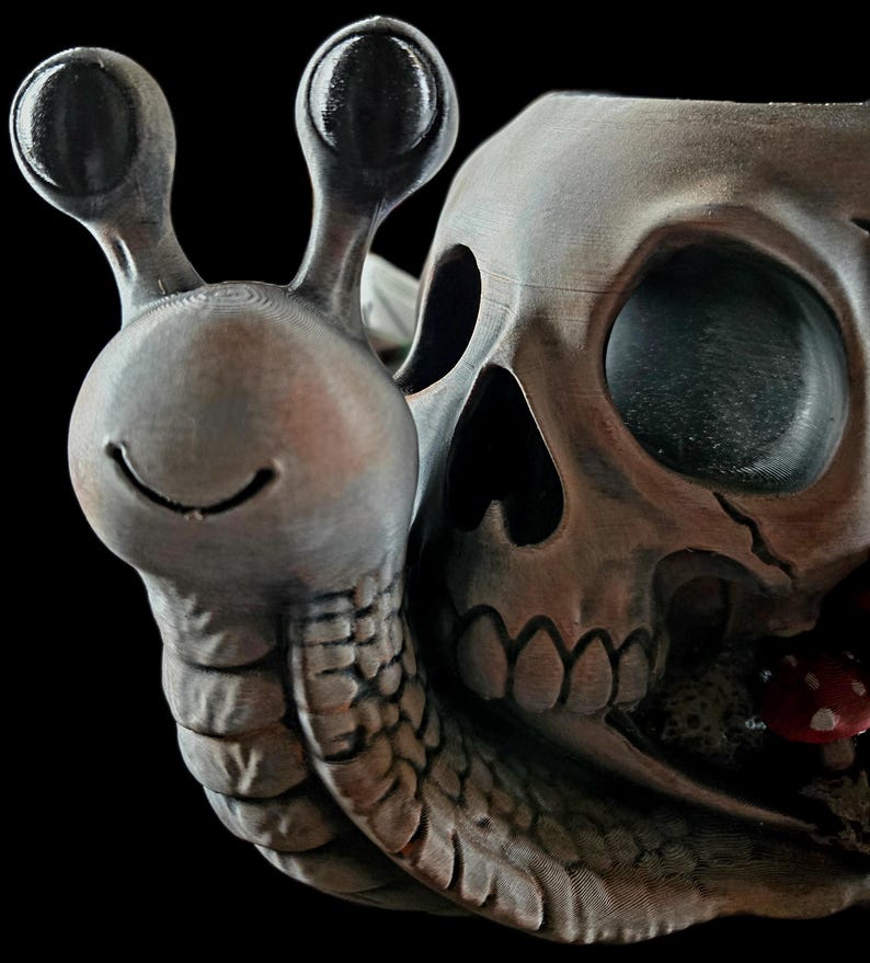 May include: A gray, textured snail figurine with a smiling face and two eye stalks. The snail shell is shaped like a human skull with hollow eye sockets and a nasal cavity. A small red mushroom with white spots is visible near the base of the skull.