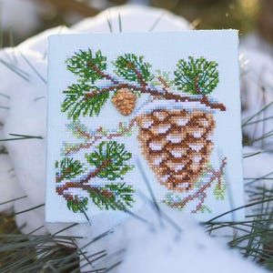May include: A cross-stitch design featuring a detailed brown pinecone and evergreen branches, dusted with snow, on a light blue background. The needlework is framed by a square, showcasing a winter or holiday theme.