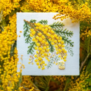 May include: Cross-stitch embroidery of yellow mimosa flowers with green leaves on a white background. The needlework is framed by a background of mimosa branches. A small, embroidered mimosa flower is in the lower right corner.