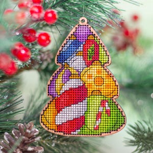 Könnte beinhalten: Weihnachtsbaumschmuck aus Holz mit Kreuzstichdesign. Der Schmuck zeigt farbenfrohe gestickte Bilder von Weihnachtskugeln und einer Zuckerstange. Der Baum hängt an einem grünen Weihnachtsbaumzweig mit roten Beeren.