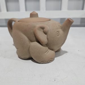 Pode incluir: Uma chaleira bege em forma de elefante. A chaleira tem uma tromba que serve de bico, uma alça e uma tampa com um pequeno botão. As características do elefante, incluindo o olho e as rugas, são sutilmente detalhadas.