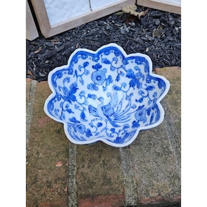 May include: A scalloped ceramic bowl featuring a white base with intricate blue floral patterns and a central phoenix illustration. The interior design displays detailed vines, leaves, and blossoms surrounding the bird, finished with a blue rim.