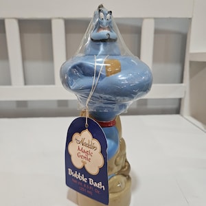 Może przedstawiać: Niebieska butelka do kąpieli bąbelkowej w kształcie Dżina z etykietą z napisem "Aladdin Magic Genie Bubble Bath". Butelka jest owinięta przezroczystym plastikiem i ma beżową podstawę. Butelka zawiera 251 ml płynu do kąpieli.