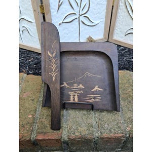 May include: A dark brown wooden crumb sweeper set consisting of a flat, square-shaped tray and a matching handheld brush. The tray features a light-colored etched landscape design of a mountain, a torii gate, and trees. The brush has a long handle.