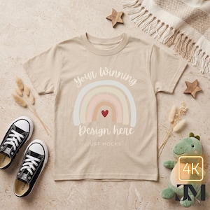 Peut inclure: Un t-shirt beige avec un motif arc-en-ciel et le texte "Your Winning Design here". Des baskets en toile noires et un dinosaure en peluche vert sont à côté du t-shirt. L'image comprend également des étoiles en bois et des éléments décoratifs.